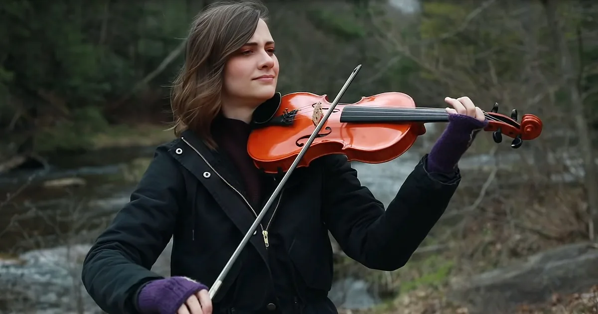 A person in a black jacket and purple gloves plays a violin outdoors near a stream.