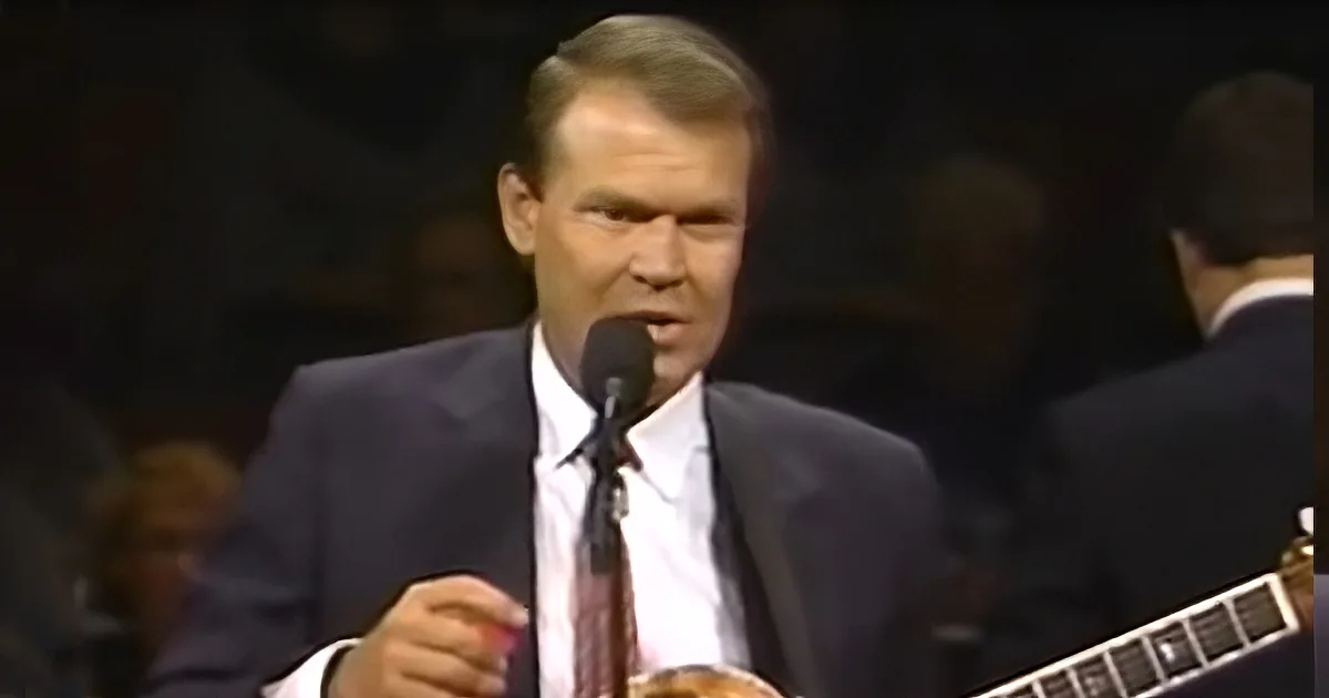 A man in a suit sings into a microphone, holding a red object, with a guitarist in the background.