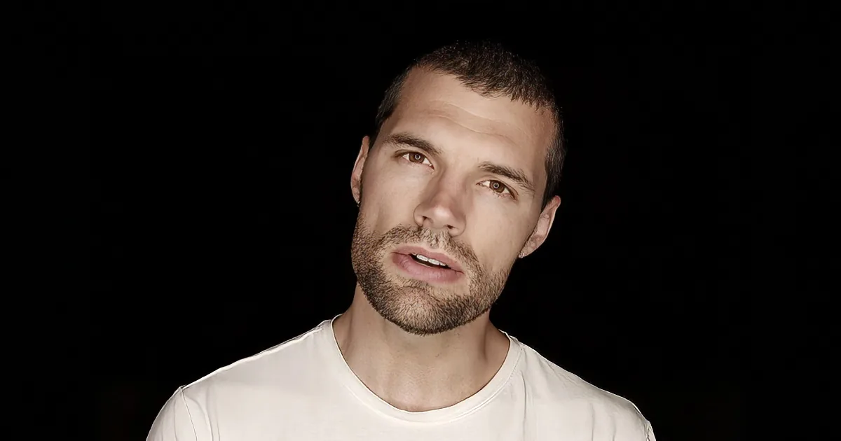 A man with short hair and a beard, wearing a white shirt, stands against a black background.