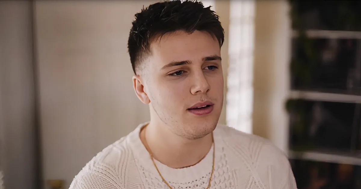 A person with short dark hair, wearing a white sweater and a gold chain, is looking to the side.