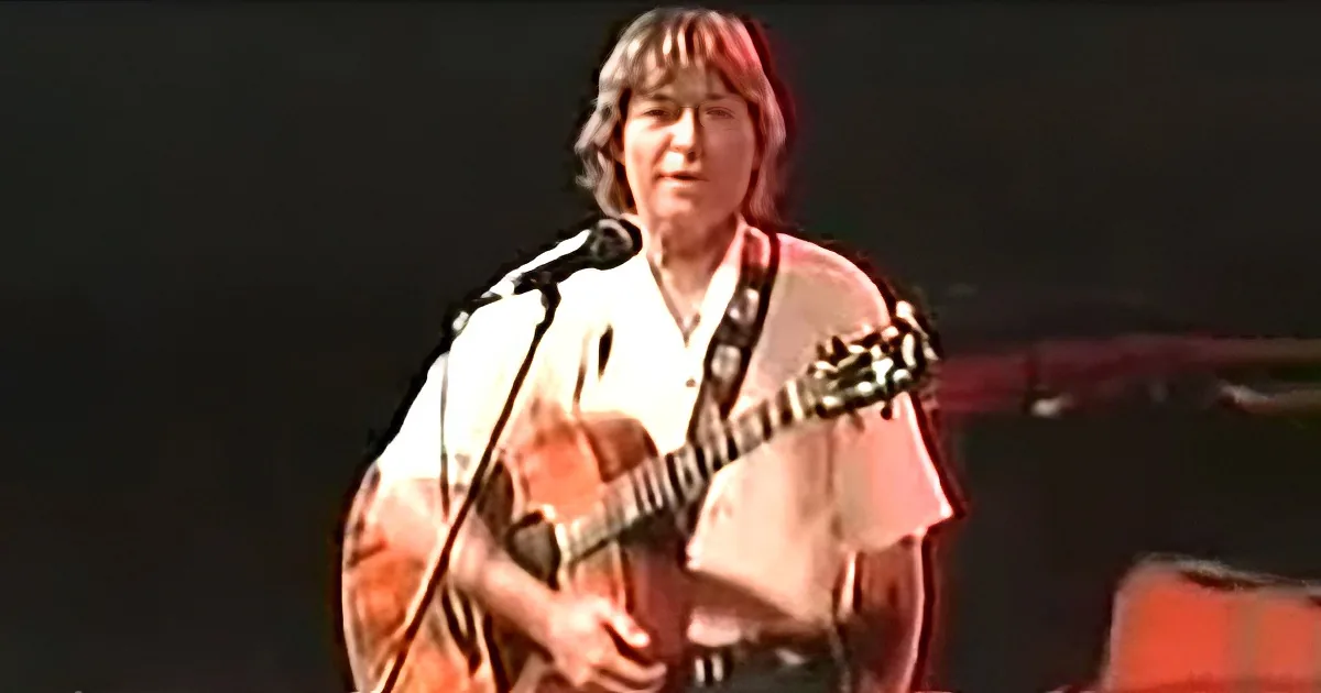 A person with shoulder-length hair plays an acoustic guitar on stage, wearing a white shirt.