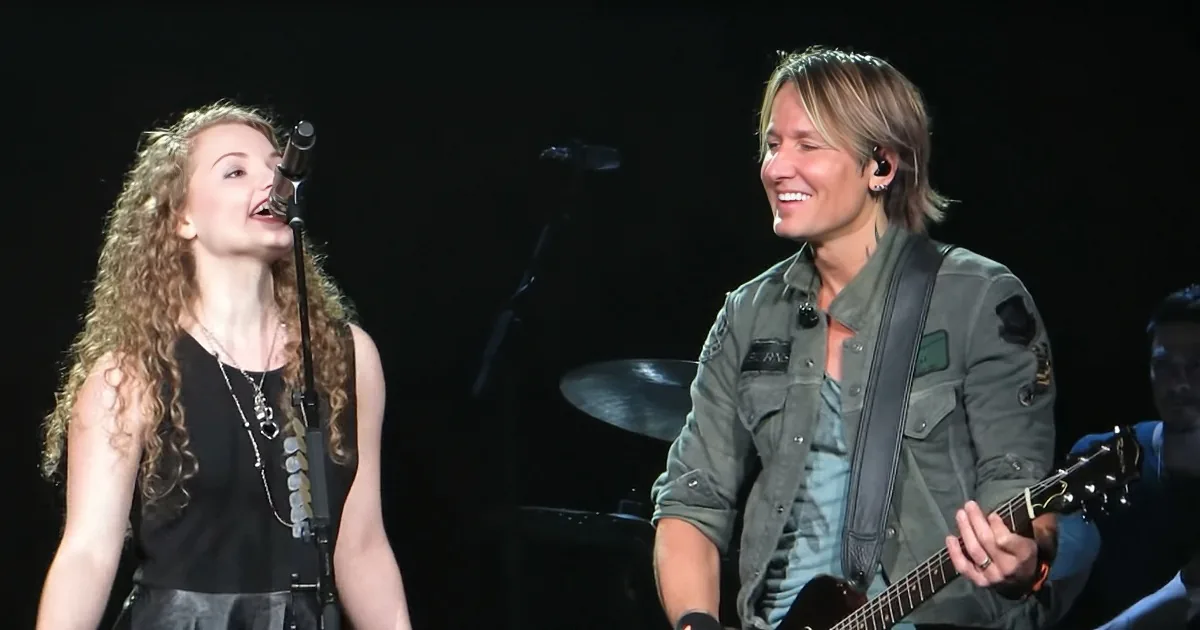 A woman sings into a microphone while a man plays guitar on stage.