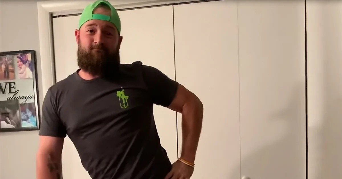 A bearded man in a green cap and black t-shirt stands indoors, hands on hips, with a framed picture on the wall behind him.