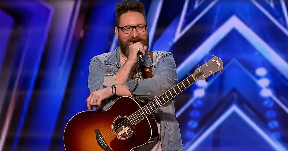 A man with a beard and glasses plays an acoustic guitar and sings into a microphone on stage.