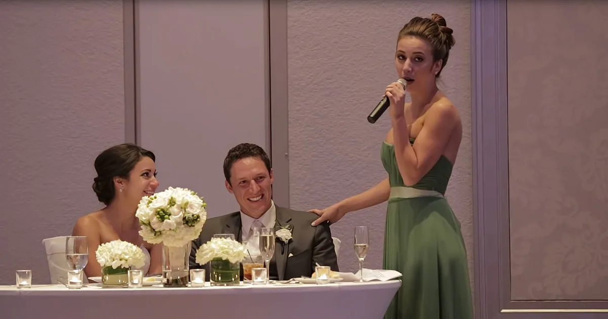 A woman in a green dress stands at a table, holding a microphone, while a couple sits smiling nearby.