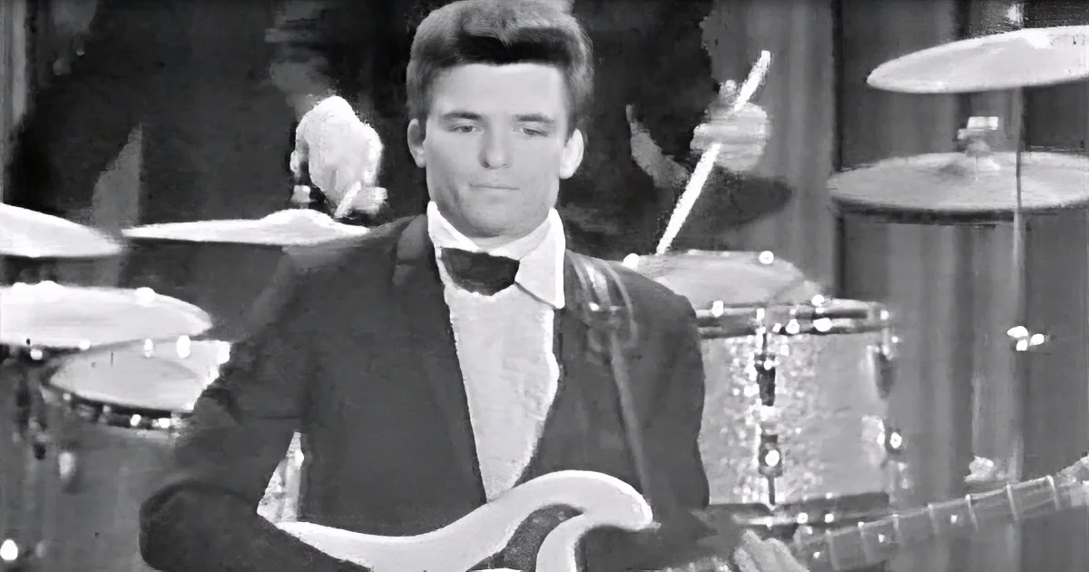 A man in a tuxedo plays a white guitar, with a drum set in the background.