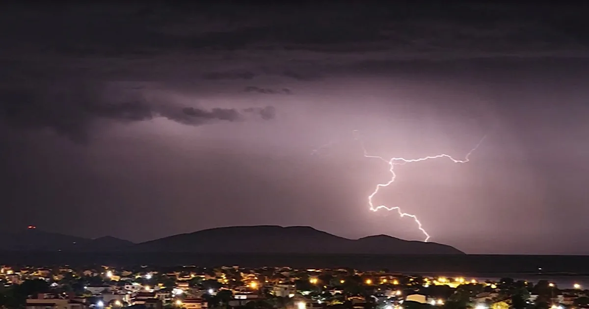 A lightning bolt strikes above a cityscape with mountains in the background under a dark, stormy sky.