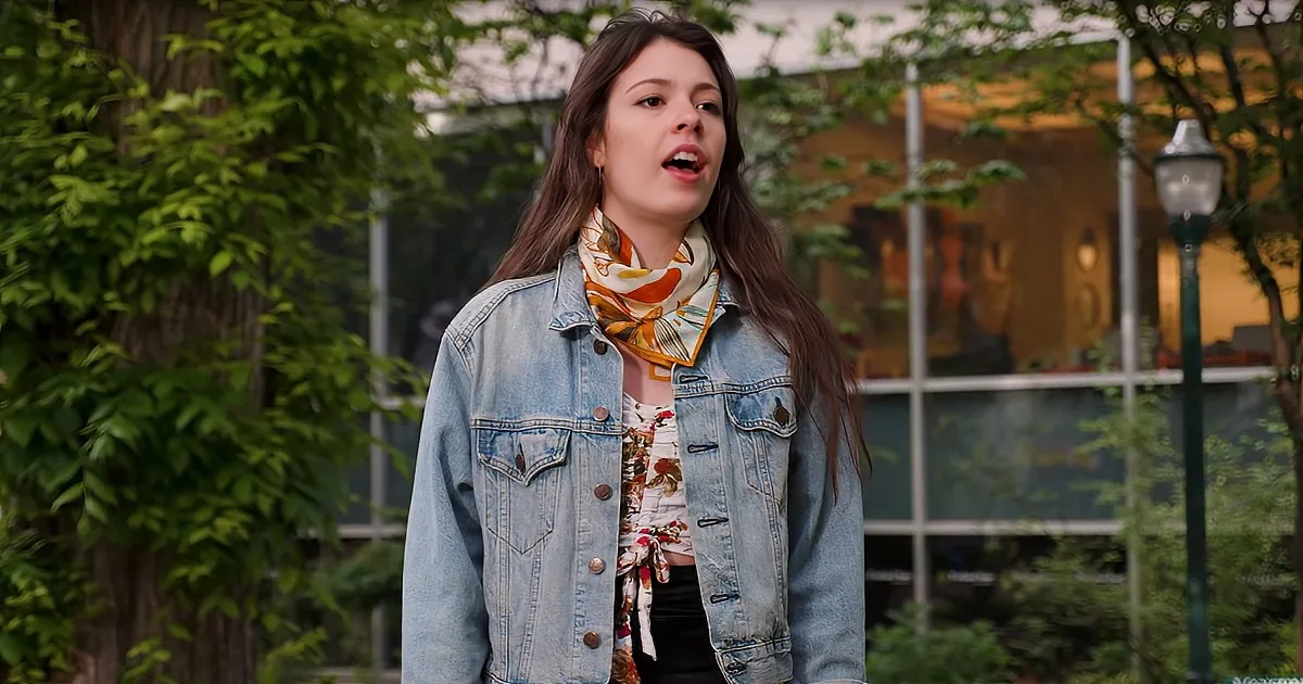 A woman in a denim jacket and floral scarf stands outdoors, surrounded by greenery and a building with large windows.