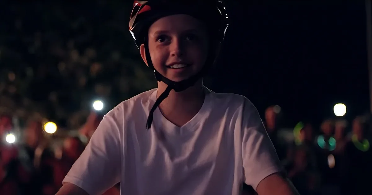 A smiling child wearing a helmet and white shirt stands in front of a crowd with glowing lights.