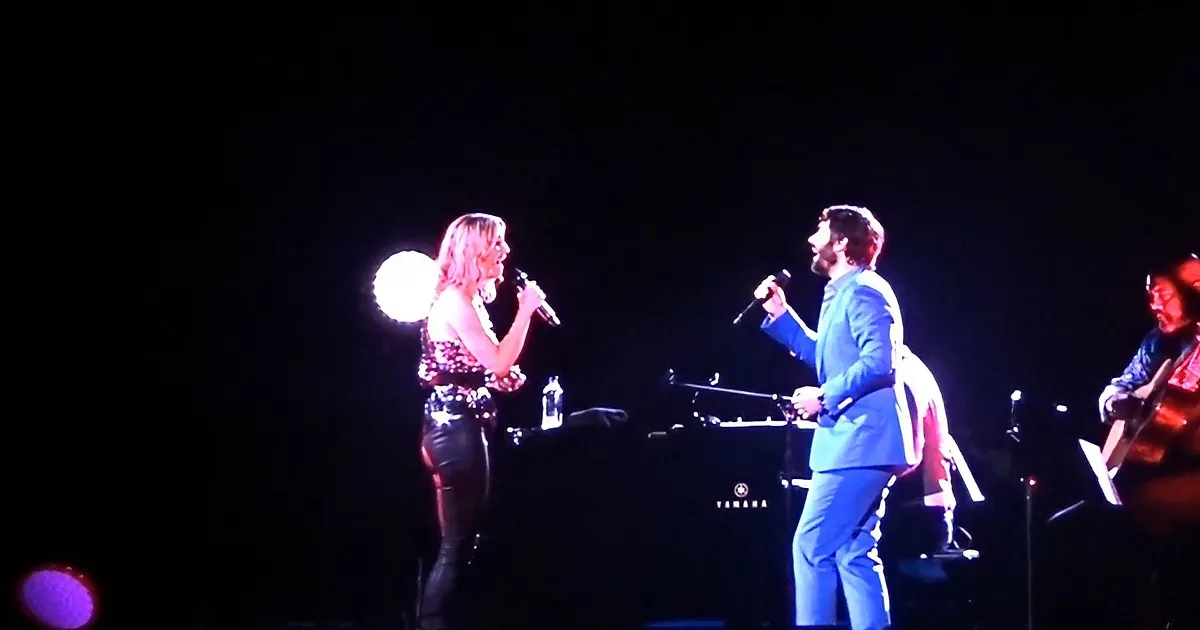 A woman in a black dress sings into a microphone, accompanied by a man in a blue suit on stage.