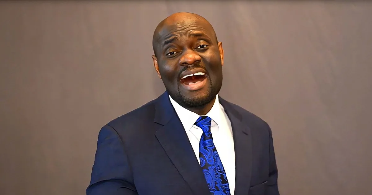 A man in a blue suit and tie is speaking with his mouth open.