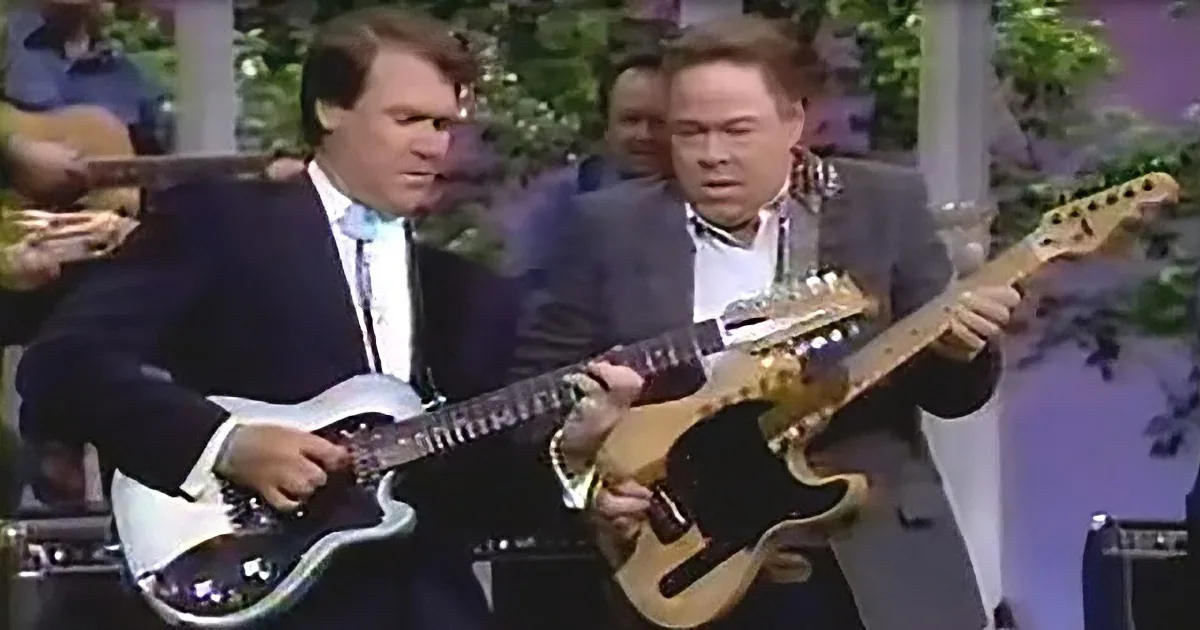 Two men in suits play guitars, one white, one brown, in a performance setting.