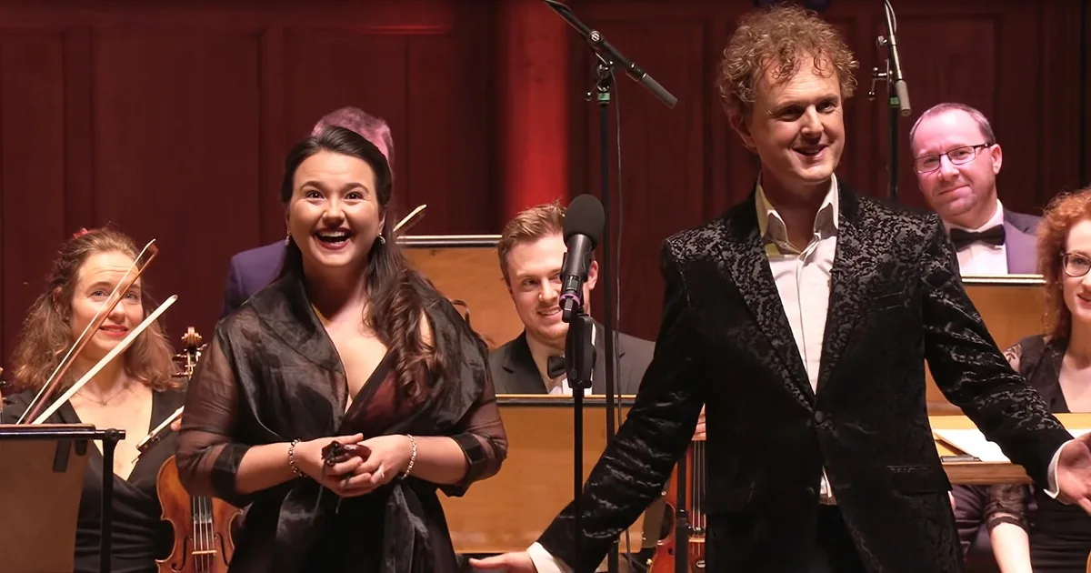 A woman in a black dress smiles, holding a microphone, with a man in a black suit beside her, both in front of an orchestra.