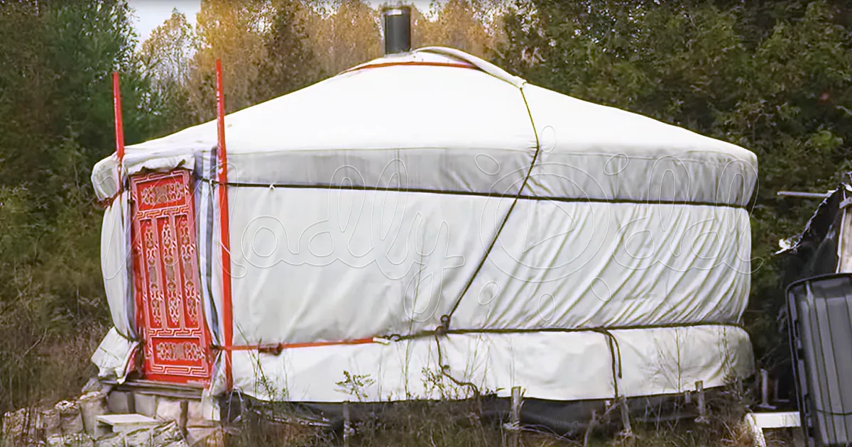 A white yurt with a red door stands amidst greenery, with a small structure visible to the right.