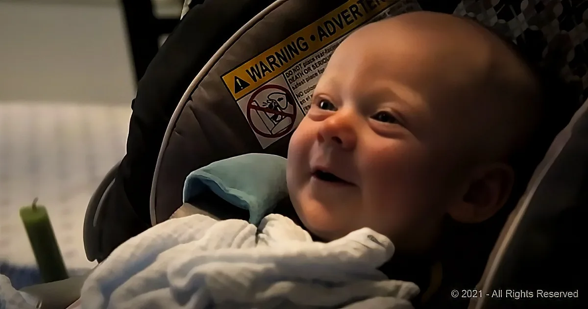 A baby in a car seat, wrapped in a blanket, with a warning label visible in the background.