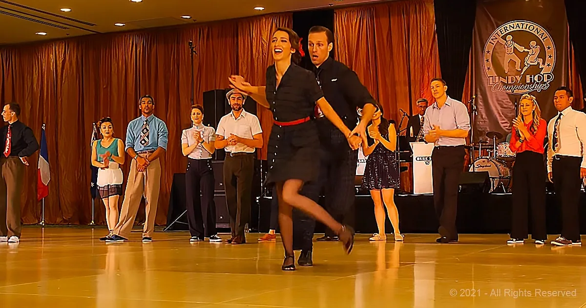 A couple dances on stage, surrounded by spectators and a banner for the Lindy Hop Championships.