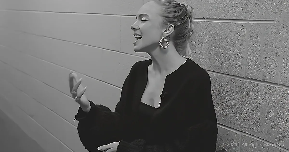 A woman in a black outfit with hoop earrings smiles and gestures with her hand against a textured wall.
