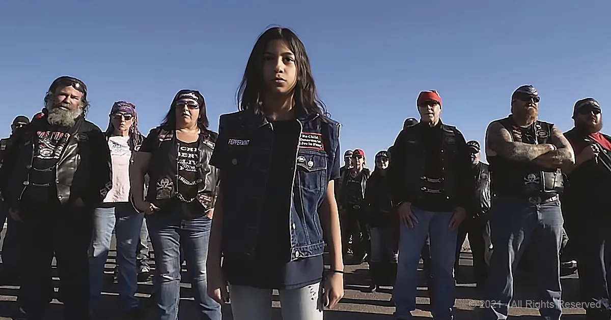 A young woman in a denim vest stands in front of a group of people wearing leather jackets and bandanas under a clear...