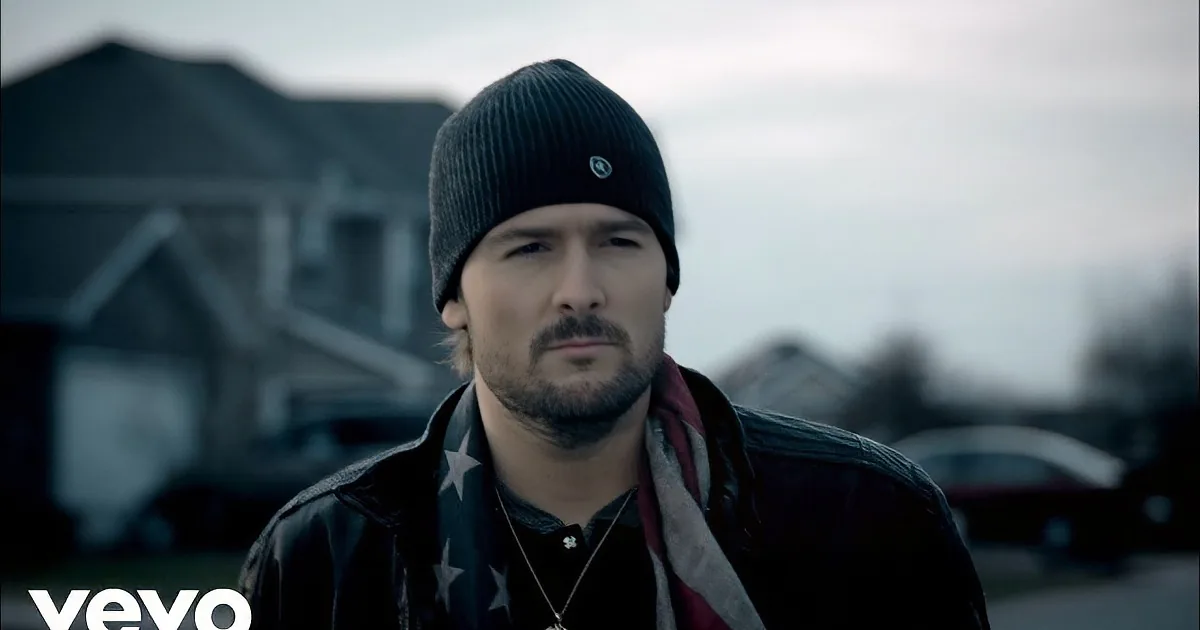 A man wearing a black beanie, leather jacket, and necklace with stars stands outdoors in a residential area.