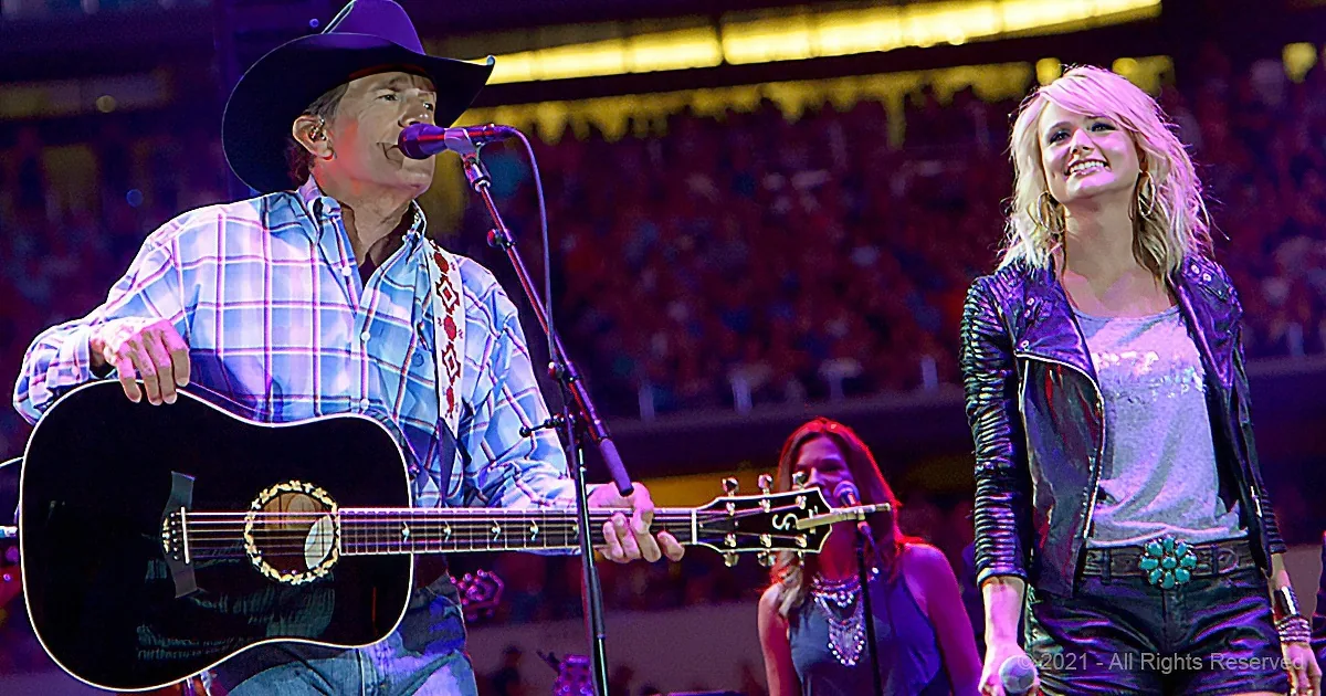 A man in a cowboy hat plays guitar while a woman in a leather jacket smiles nearby at a concert.