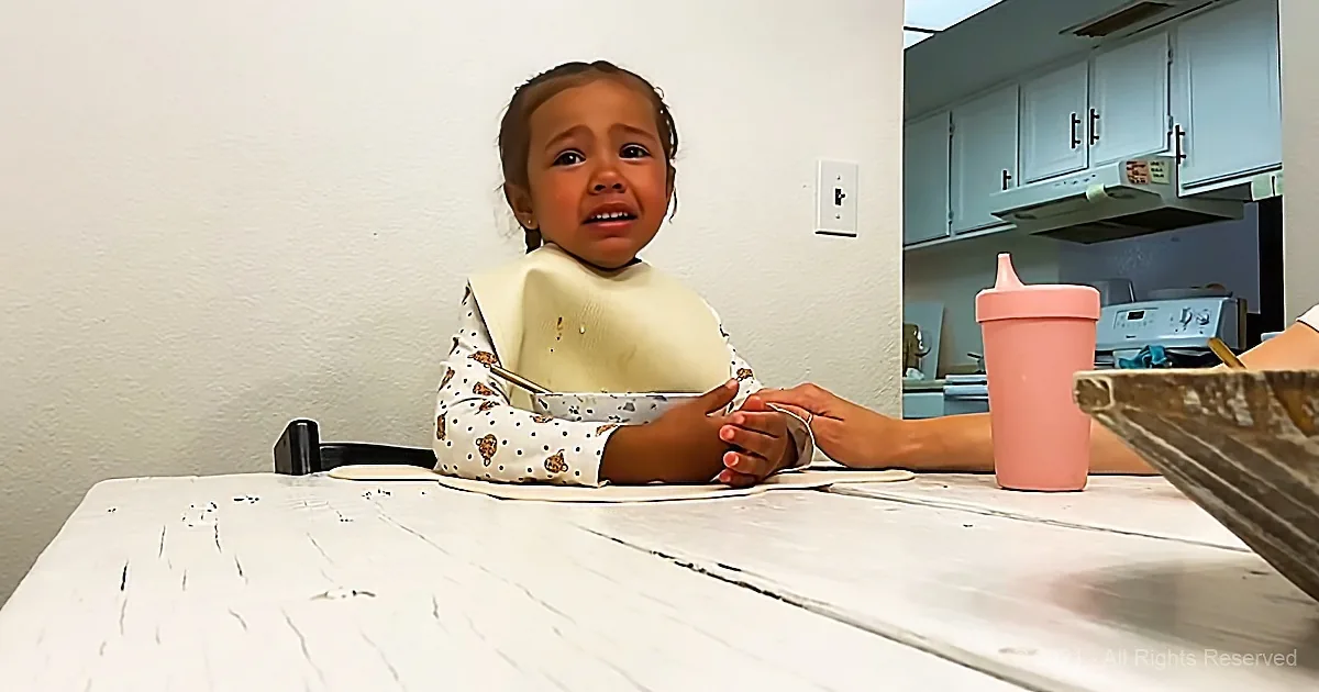 A young child wearing a bib sits at a table, holding hands with an adult, with a pink sippy cup nearby.