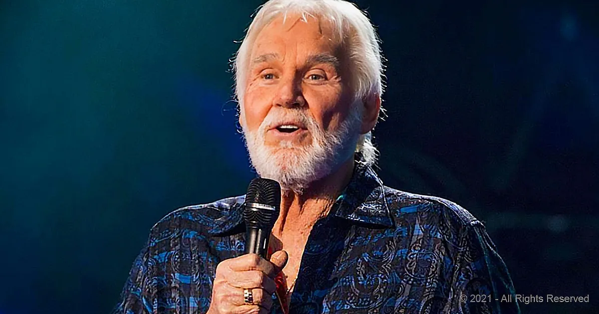 A man with white hair and a beard holds a microphone, wearing a patterned shirt, against a dark background.