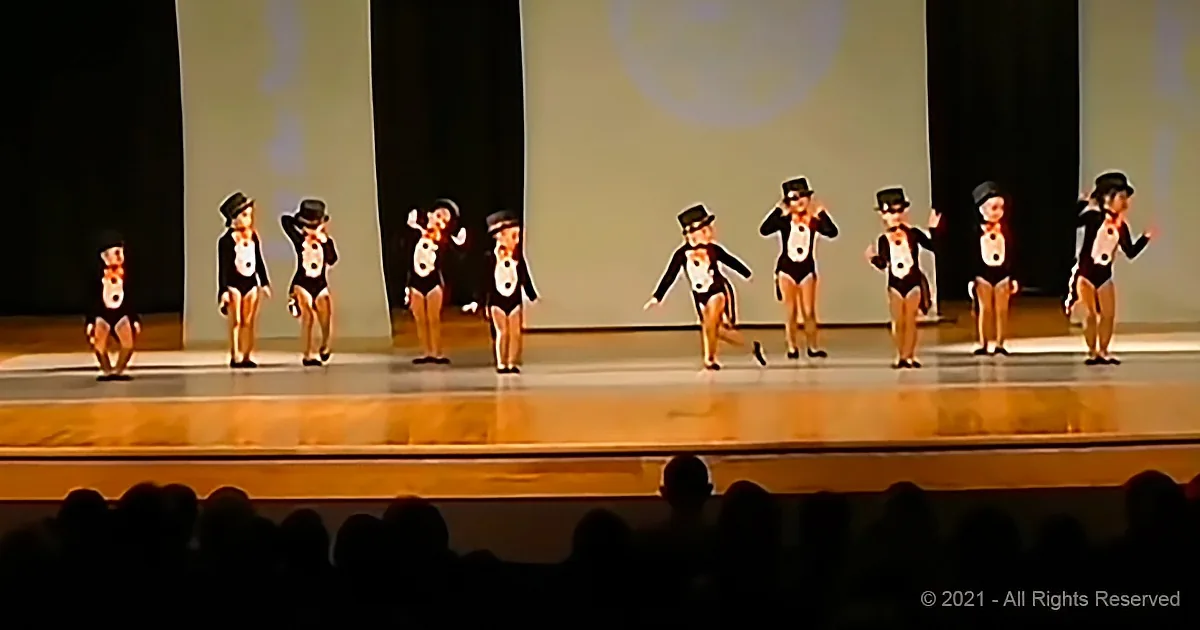 Children in black outfits and hats perform a synchronized dance on a stage with a bright background.