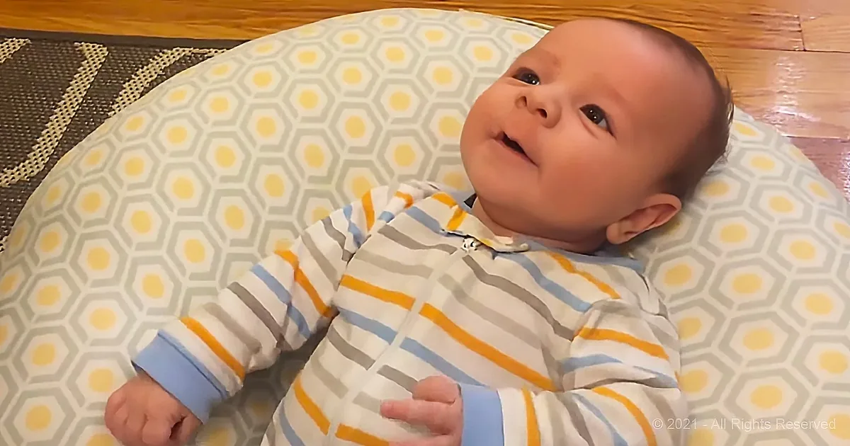 A baby in striped pajamas lies on a hexagonal-patterned pillow, looking up with a curious expression.