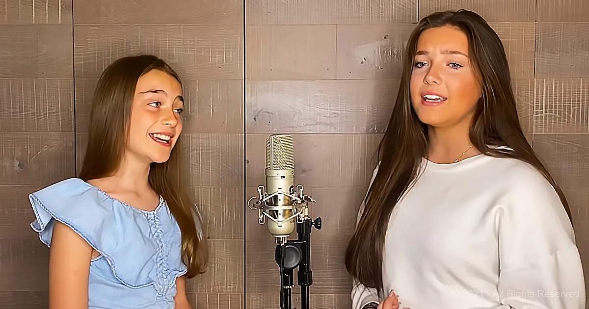 Two individuals sing in front of a microphone, one smiling, the other speaking, in a studio setting.