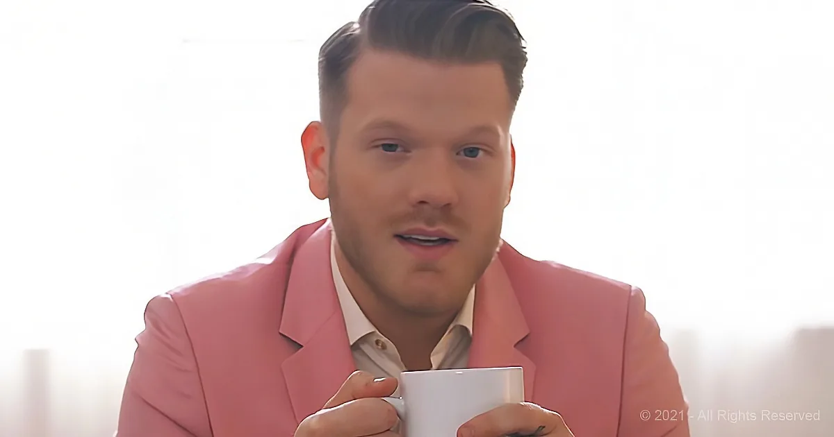 A man in a pink suit holds a white mug, looking slightly to the side.