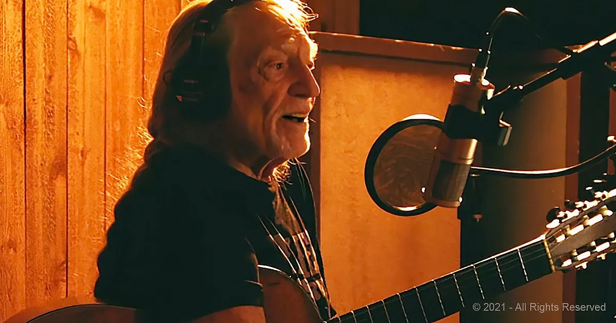 An older man wearing headphones sings into a microphone while playing an acoustic guitar in a warmly lit studio.