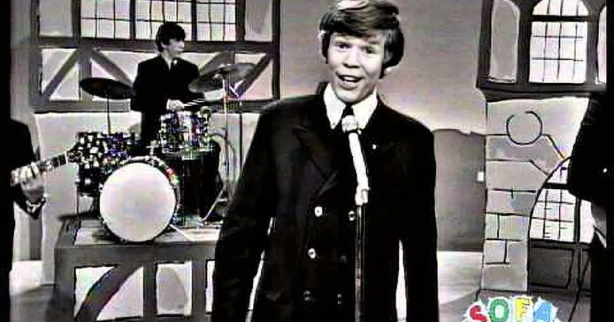 A man in a suit sings into a microphone, with a drummer and a window in the background.