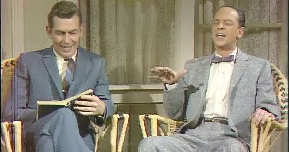 Two men in suits are seated, one gesturing while the other holds a book, appearing engaged in a discussion.