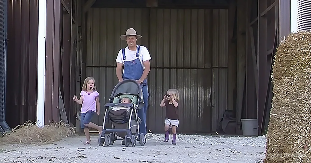 A man in overalls pushes a stroller with a baby, accompanied by two children, near a barn.