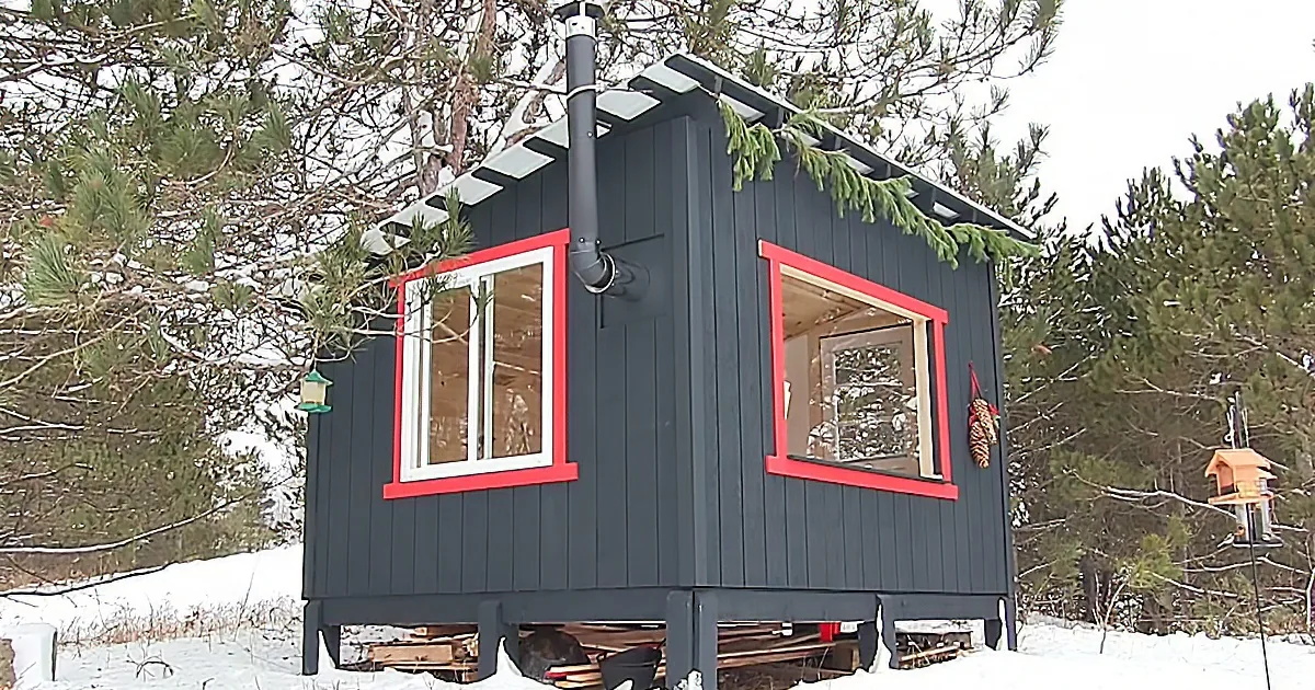 A small, dark gray cabin with red-framed windows sits on stilts in a snowy forest.