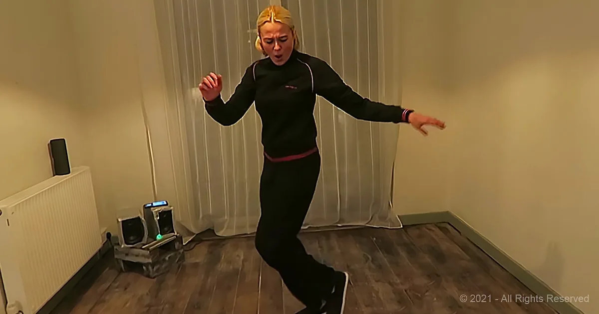 A person in a black outfit is dancing energetically in a room with wooden floors and a radiator.