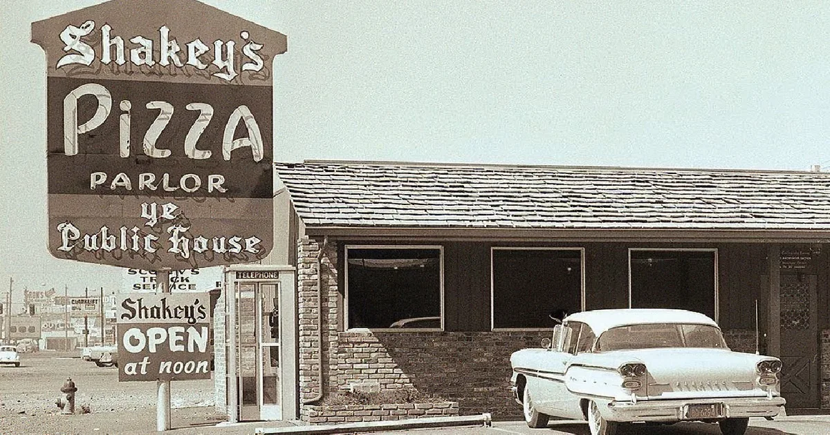 A man in a white shirt and tie holds two large pizzas in a restaurant setting.
