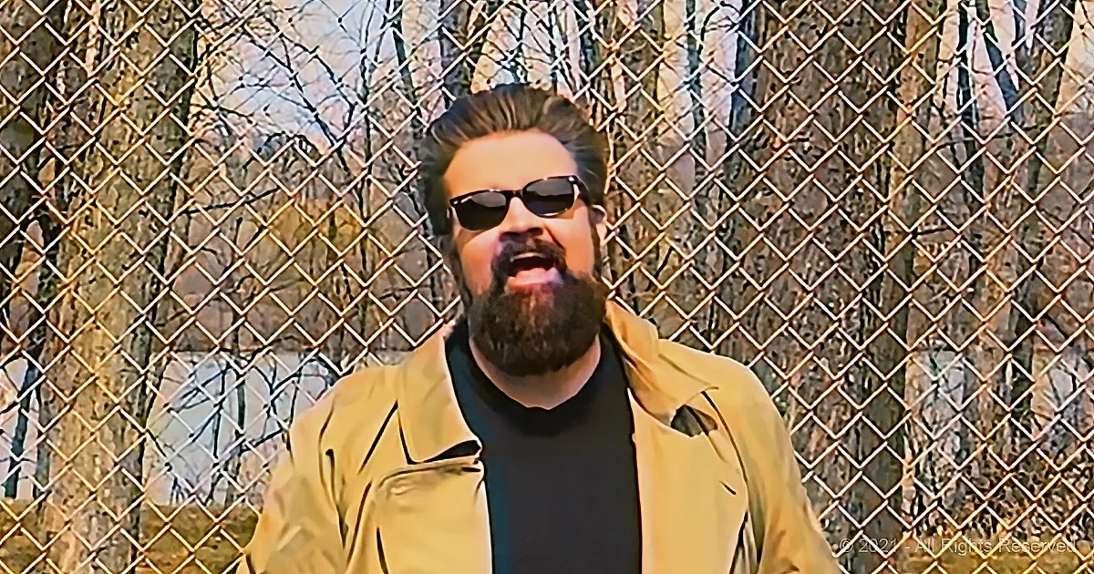 A man with a beard and sunglasses stands in front of a chain-link fence, wearing a yellow jacket.