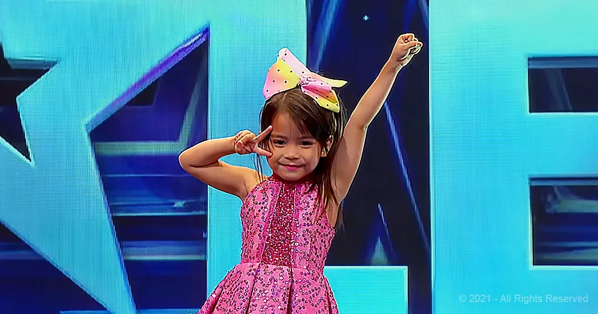 A young girl in a pink sequined dress and colorful bow on her head raises her arms and makes a peace sign.