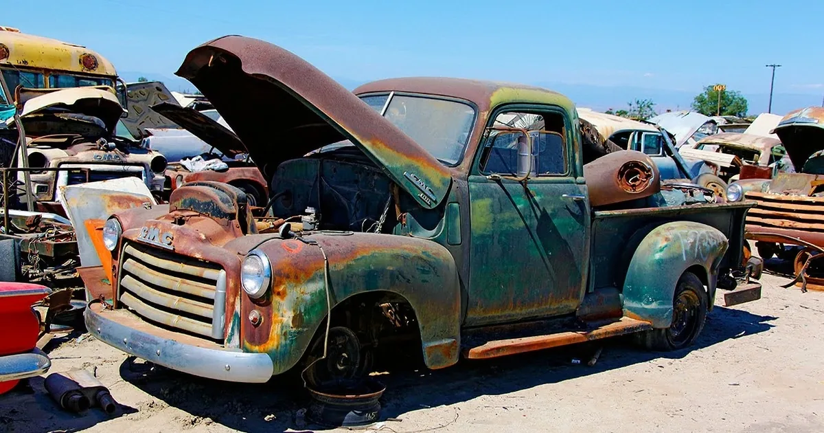 An old, rusted GMC truck with its hood open sits in a junkyard under a bright blue sky.