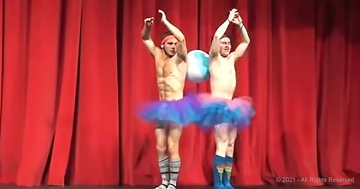 Two shirtless men in tutus perform on stage with a red curtain backdrop.