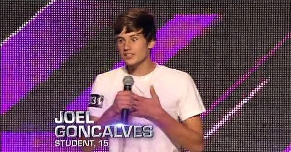 A young man in a white t-shirt holds a microphone on stage with a vibrant pink and purple background.