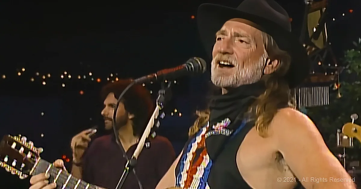 A man with long hair and a beard, wearing a hat and a black tank top, is singing and playing a guitar on stage.