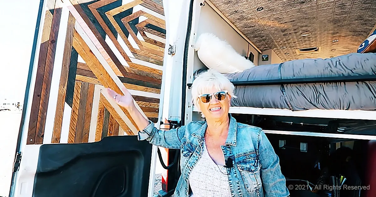 A smiling woman in a denim jacket stands inside a van, gesturing towards a wooden structure.