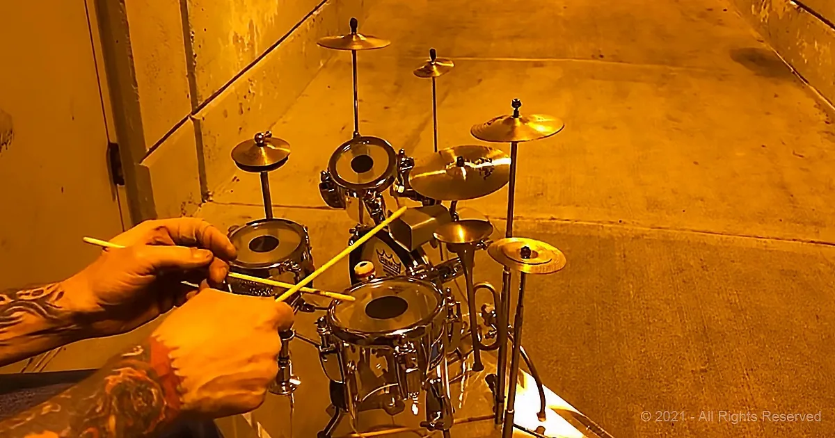 A person with tattooed hands holds drumsticks, preparing to play a drum set in a dimly lit room.
