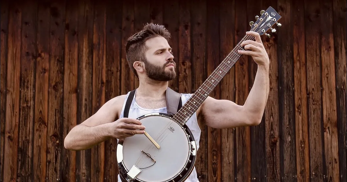 A man with a beard plays a banjo against a wooden fence.