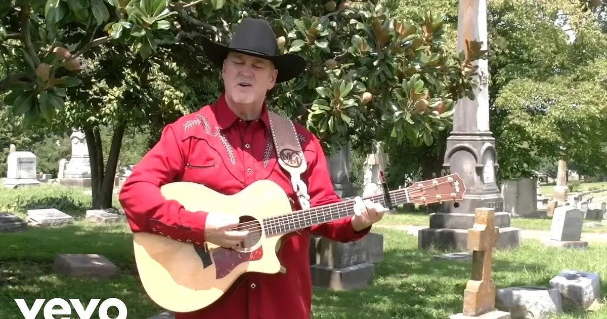 A man in a cowboy hat and shirt sings passionately while playing a white guitar in a cemetery.