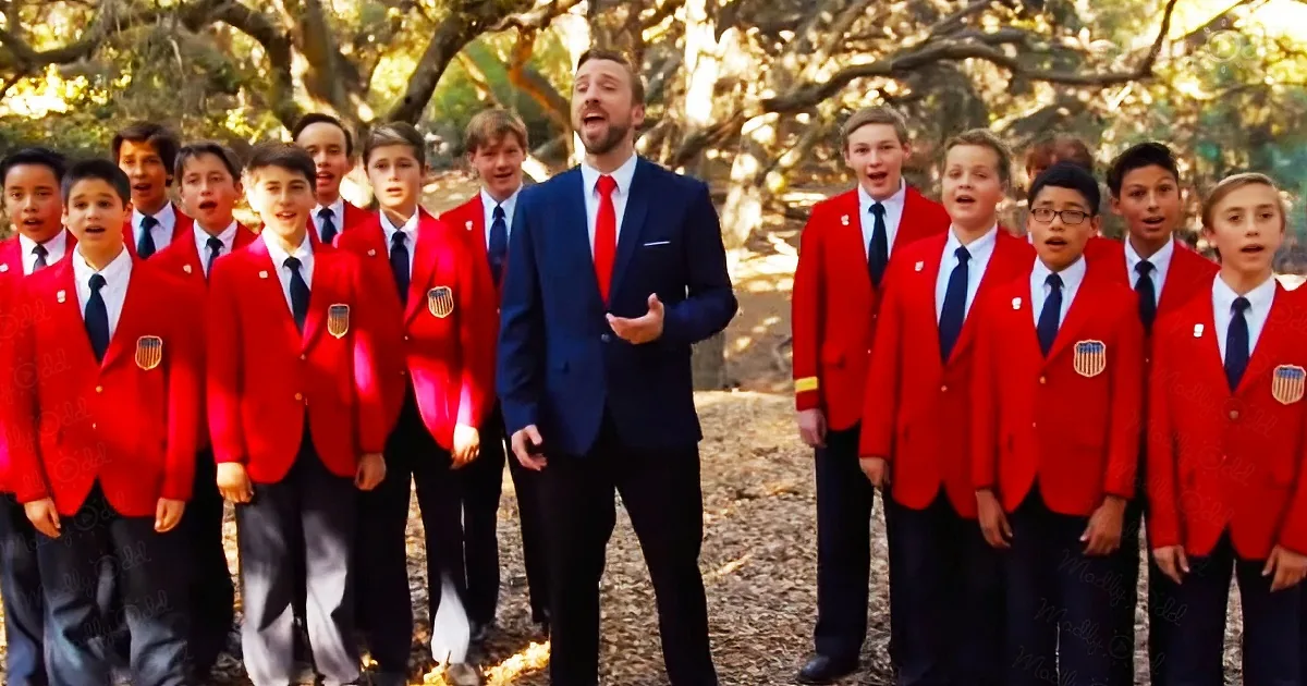 A man in a blue suit leads a group of boys in red jackets singing outdoors.