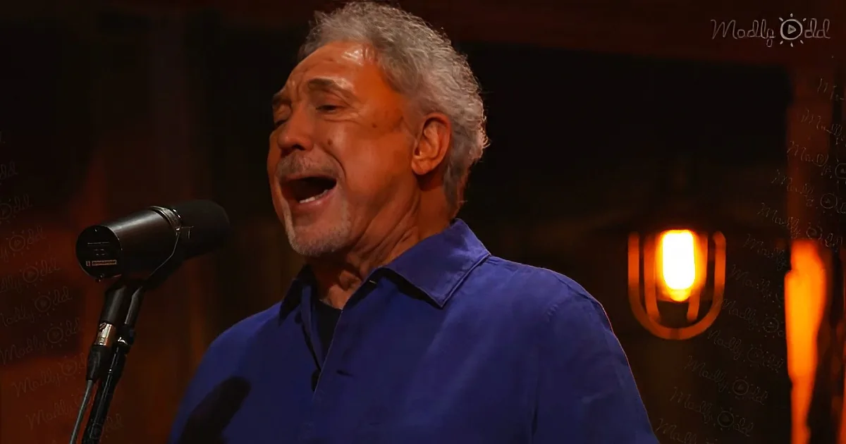 A man in a blue shirt sings into a microphone, with a warm light in the background.