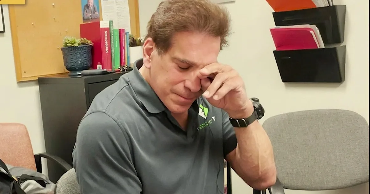 A man in a green shirt sits at a desk, holding his hand to his face, with a bookshelf and plants in the background.